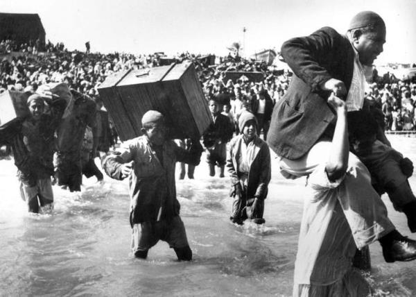 A man carrying luggage wades through water while another lifts an elderly man on his shoulders.