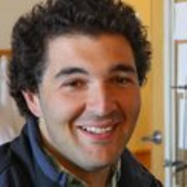 Adam Oken, B.A., sits at a computer smiling. He has short dark curly hair and a blue jacket.